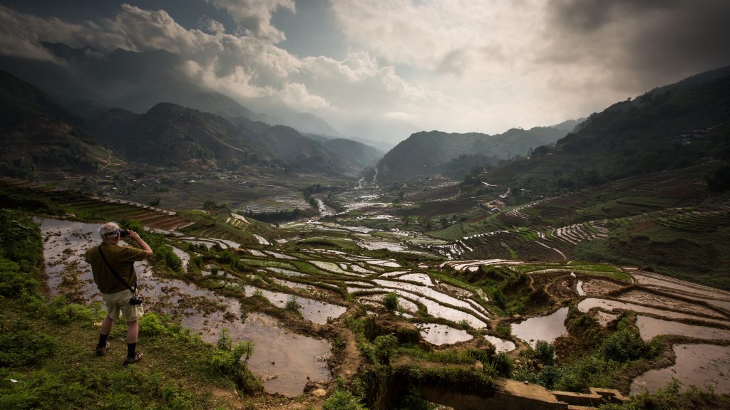 A man with a camer is taking in the view and capturing an incredible rice terrace landscape from above, with paddies filled with water and a view of the whole valley.